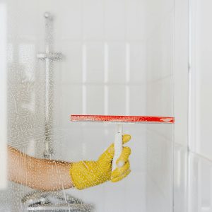 A person wearing a yellow glove cleans a shower glass with a squeegee, emphasizing hygiene and cleanliness.